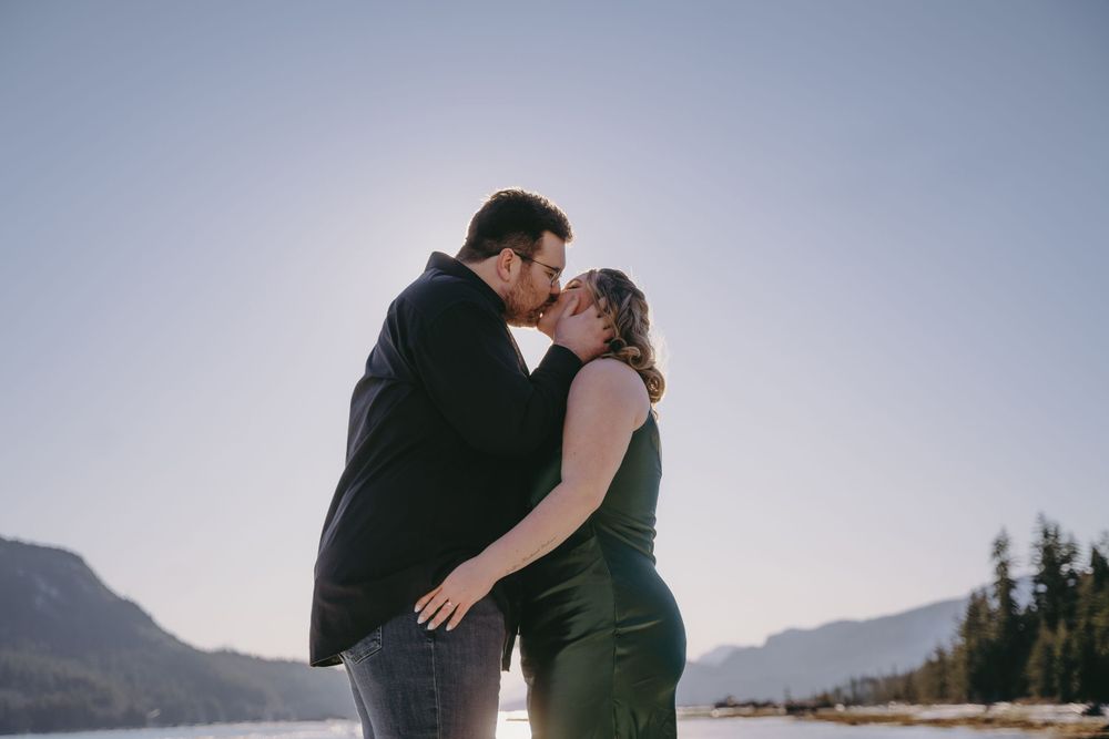 A romantic couple embraces during sunset at a mountain lake with evergreen trees and mountains in the background.