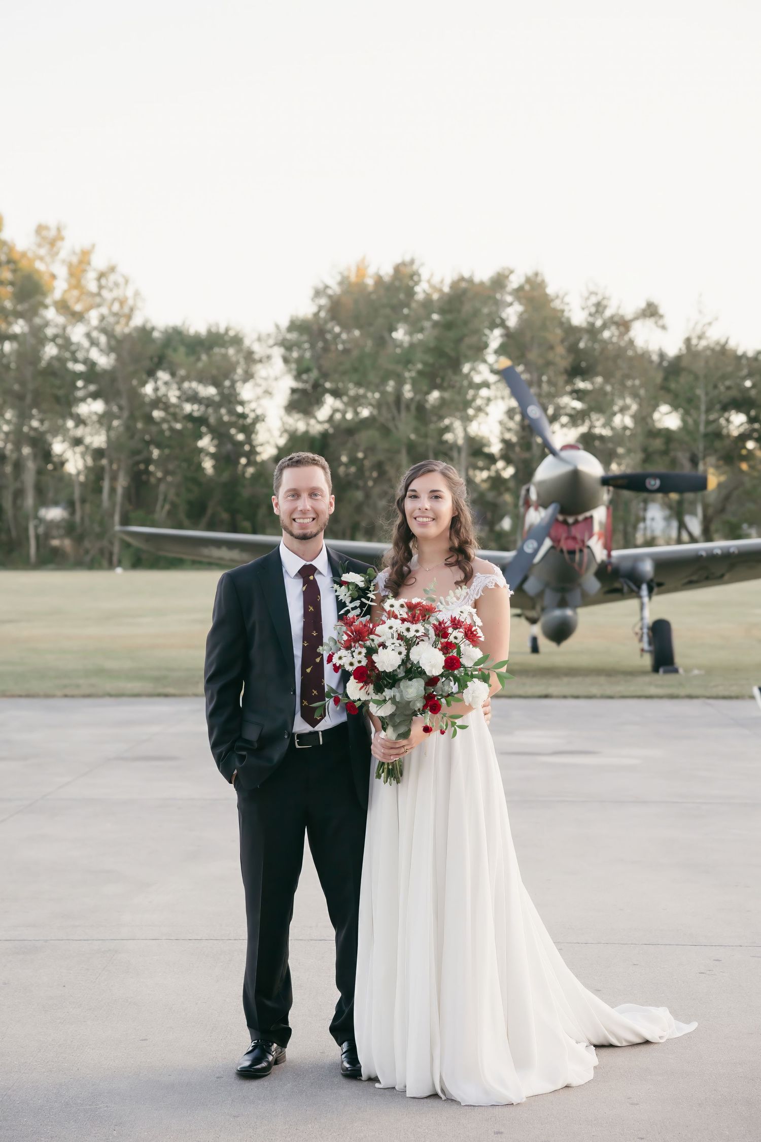 A couple stands together at an outdoor venue with a beautiful floral bouquet and trees in the background.