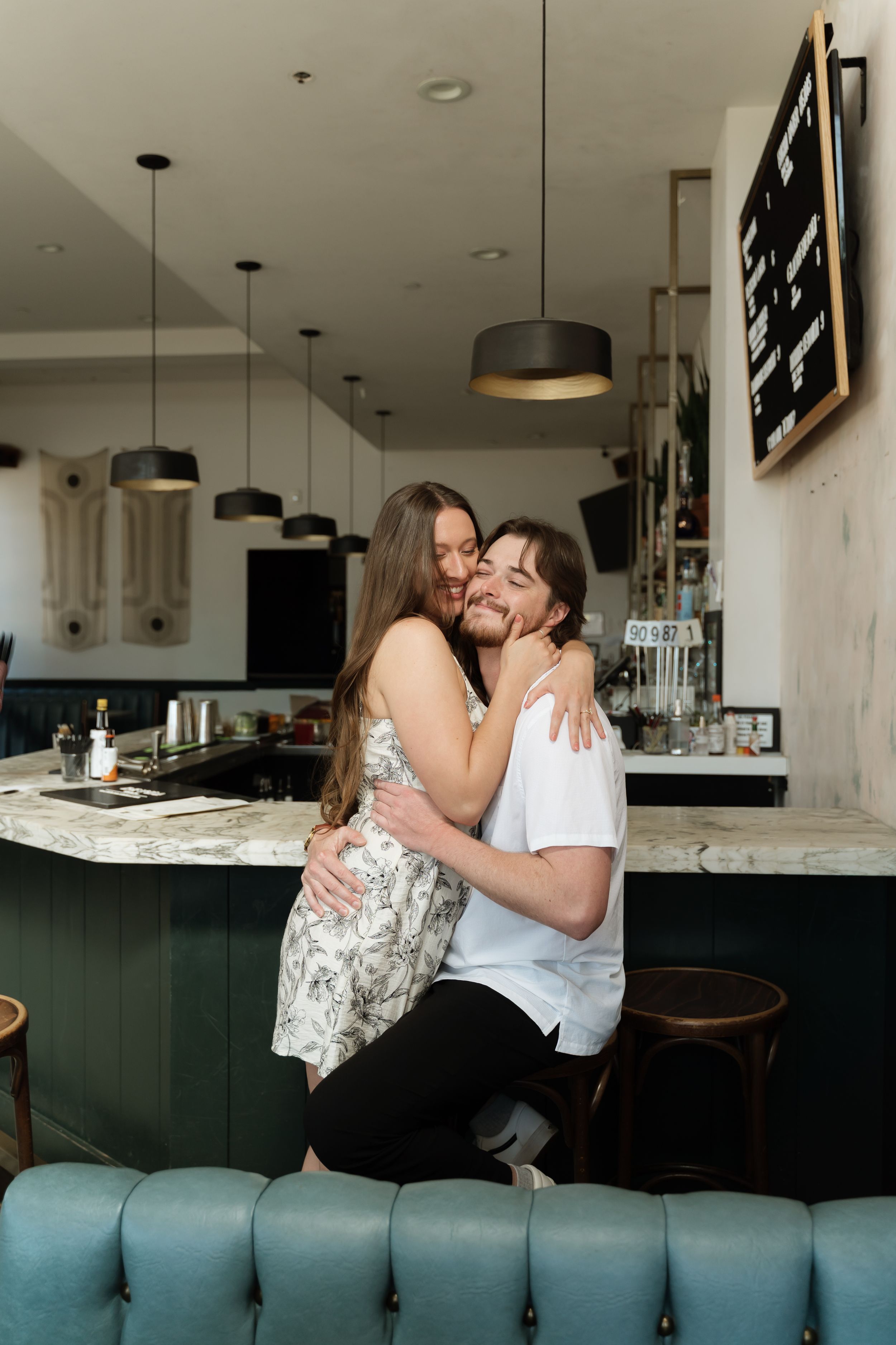 Couple embracing and smiling at a bar counter, woman in floral dress hugging man in white t-shirt on a barstool.