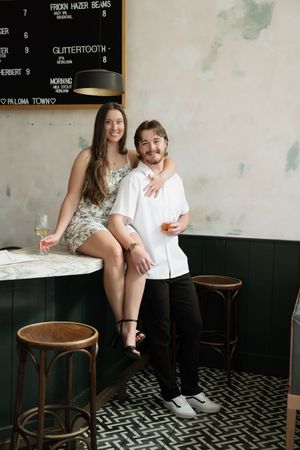 Couple smiling and holding drinks at a stylish bar with green paneling, marble counter, and wooden stools.