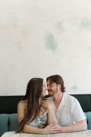 A couple shares a tender moment, smiling at each other while seated at a marble table against a soft watercolor wall.