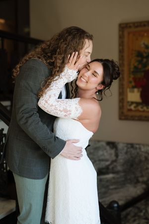 A groom in a gray suit tenderly kisses his smiling bride in a white lace wedding dress during an intimate indoor moment.