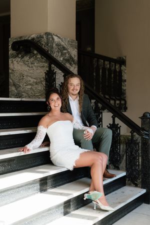 Bride in white lace off-shoulder dress and groom in gray suit sitting on marble stairs smiling.