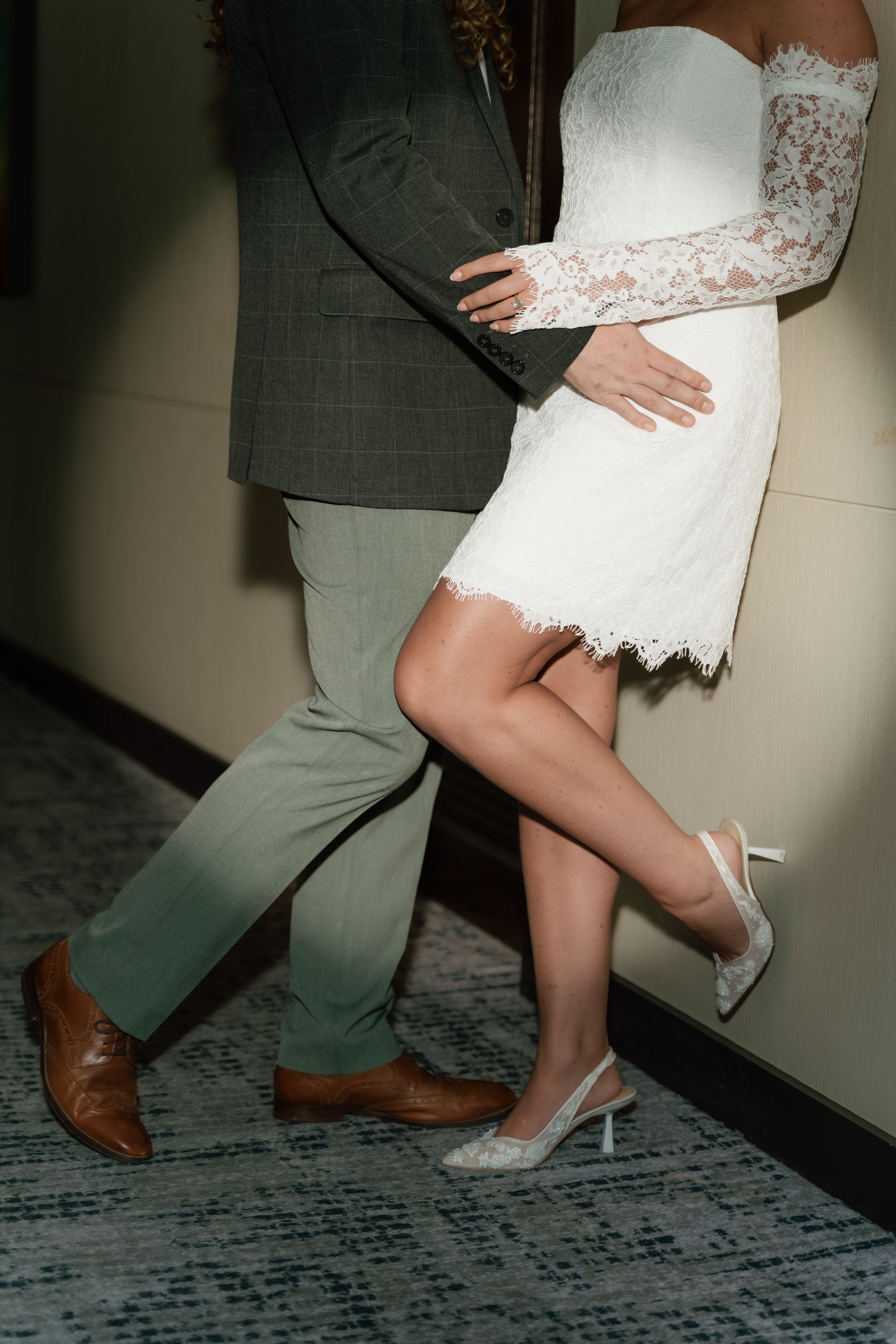 Bride in white lace mini dress and groom in sage suit embrace in a romantic pose, showing elegant wedding attire.