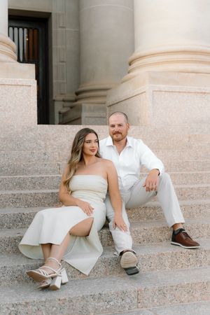Couple in white outfits sitting on stone steps of a classical building for an elegant engagement photo session.