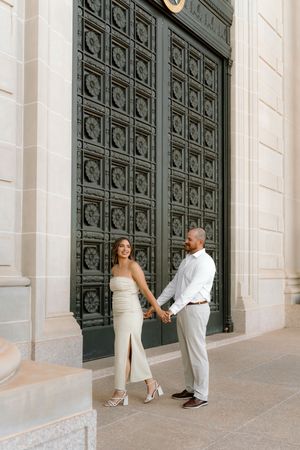 Couple holding hands in front of ornate dark green doors, woman in cream dress, man in white shirt and gray pants.