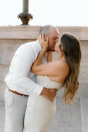 Couple sharing a romantic kiss outdoors, dressed in elegant white outfits, embracing tenderly near a stone wall.