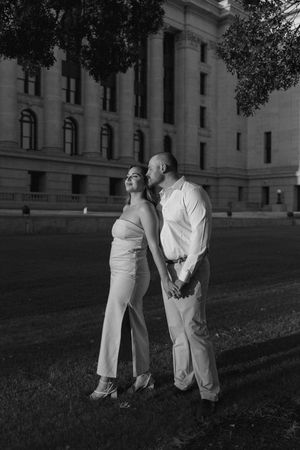 Black and white photo of a couple standing together at night in front of a grand building, man kissing womans cheek.
