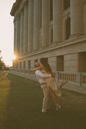 Couple embracing at sunset beside a grand neoclassical building with tall columns, woman lifted off ground.