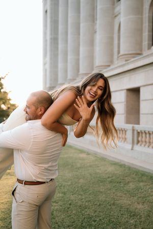 A joyful couple celebrates their engagement outdoors, woman showing ring while being lifted by man near a grand building.