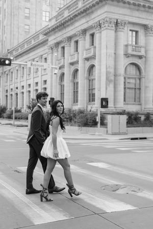 Black and white photo of an elegant couple crossing a city street, woman in white dress, man in suit, classic building backdrop.