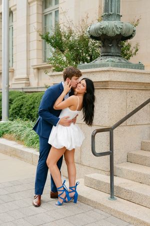 Couple embracing near stone steps, man in blue suit kissing woman in white mini dress and blue heels.