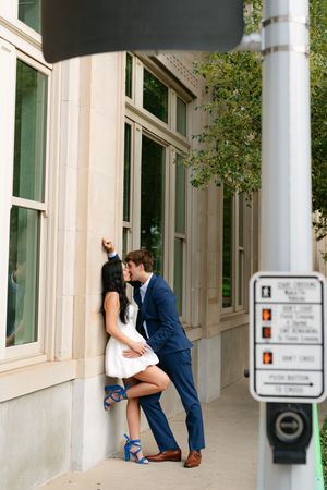 Couple sharing a romantic kiss against a limestone building wall, woman in white dress and blue heels.