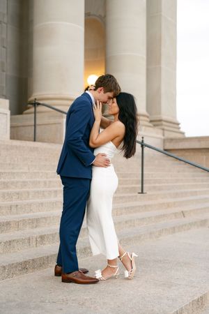 Couple sharing a romantic kiss on grand stone steps, man in blue suit and woman in white jumpsuit.