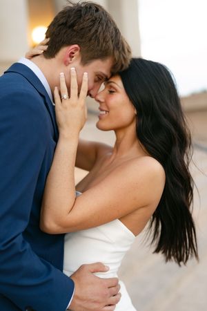 A bride in a white dress and groom in a blue suit share a romantic forehead touch, showcasing her diamond engagement ring.