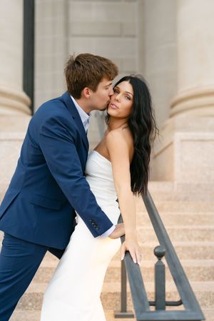 Groom in blue suit kisses bride in white dress on outdoor staircase.