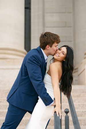 Man in blue suit kissing smiling woman in white dress on outdoor stone staircase.