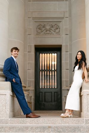 Couple posing apart on stone steps before ornate door, man in blue suit and woman in white dress.