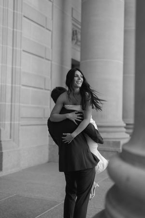 Black and white photo of a man lifting a smiling woman near elegant stone columns in an urban setting.
