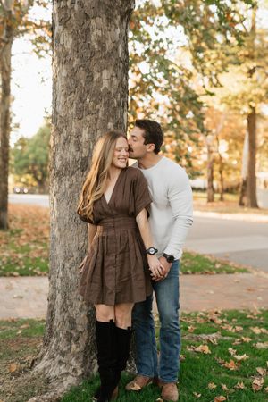 Couple sharing a romantic kiss by a tree in autumn park, woman in brown dress and knee-high boots.