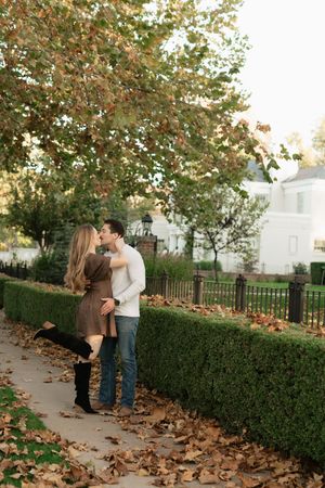 Couple sharing a romantic kiss on a leaf-covered sidewalk surrounded by autumn trees and manicured hedges.