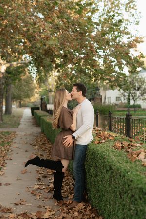 Couple sharing a romantic moment on a leaf-covered sidewalk in autumn, surrounded by colorful trees and a green hedge.