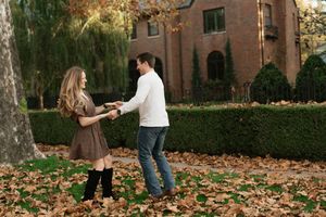 Couple holding hands and dancing among autumn leaves outside a brick building surrounded by green hedges.