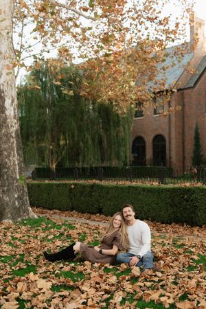 Couple sitting together on autumn leaves under a tree in a garden with a brick building backdrop.