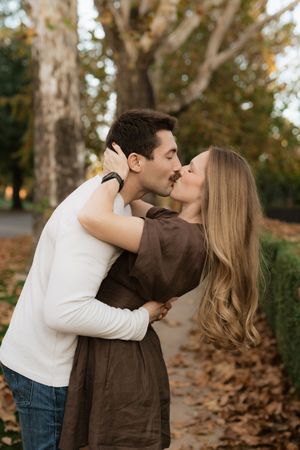 Couple sharing a romantic kiss in a park surrounded by fallen autumn leaves and trees.