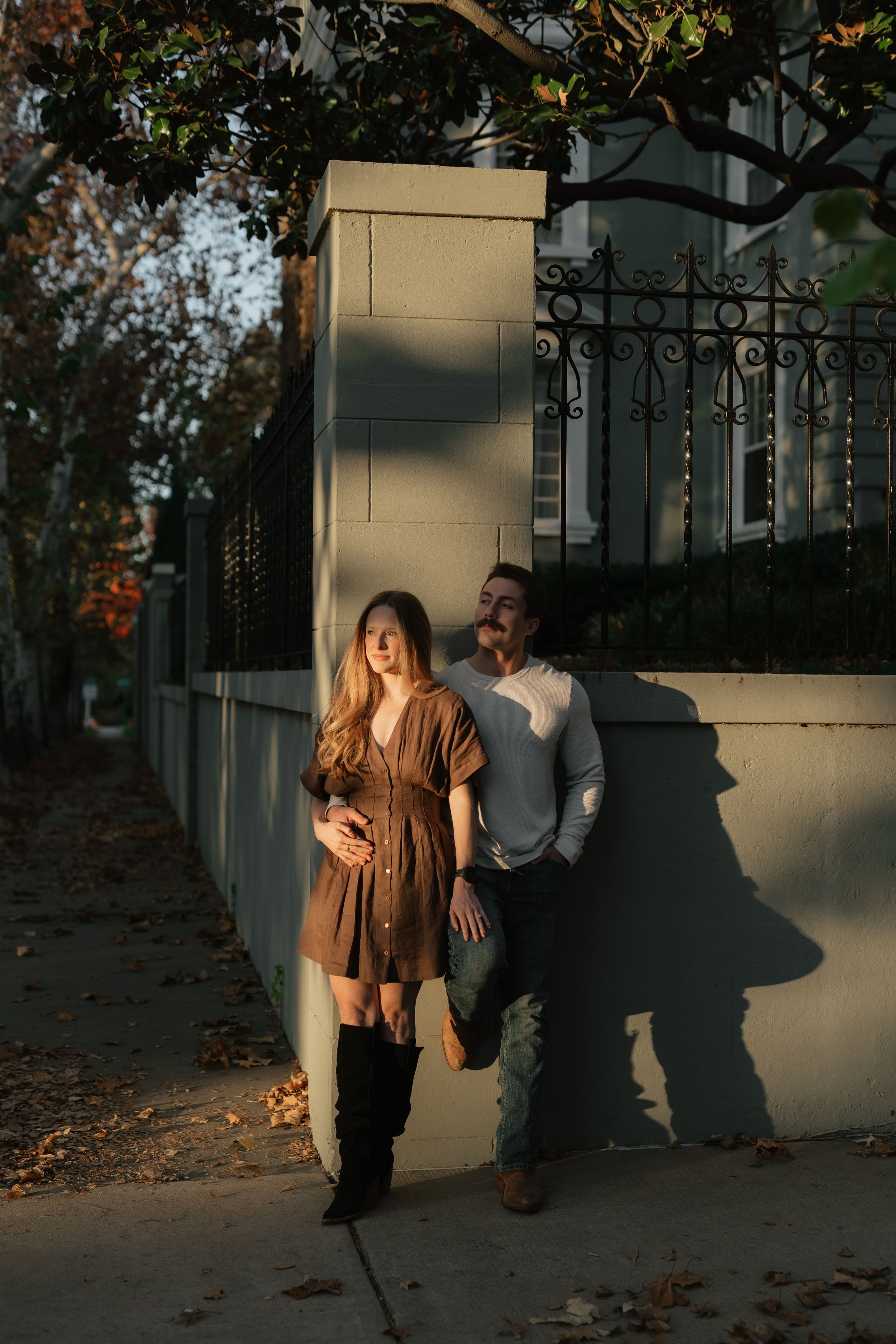 Pregnant couple leaning against a sunlit wall on a sidewalk in autumn, woman in brown dress and knee-high boots.