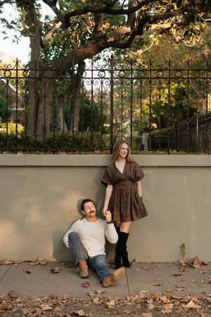 Couple holding hands by iron fence in fall, man sitting on ground, woman in brown dress and black boots.