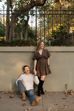 Couple holding hands outdoors, man sitting against wall, woman in brown dress and black boots smiling in autumn park.