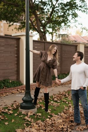 Woman in brown dress and boots playfully holds a lamp post while holding hands with smiling man among fall leaves.