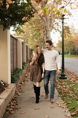 Couple walking together on a leaf-covered sidewalk in autumn, smiling and laughing in casual fall outfits.