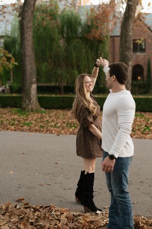 Happy couple dancing outdoors in autumn park, woman in brown dress and black boots being twirled by man in white sweater.