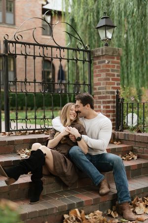 Couple sitting on brick steps by an ornate iron gate, holding hands and smiling in autumn setting.