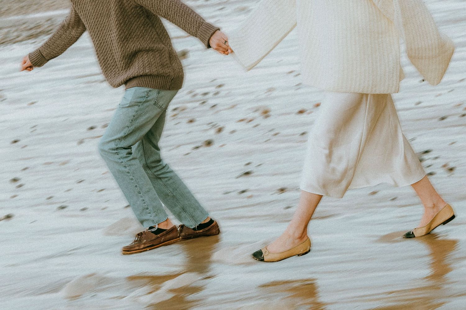 Two people walking barefoot on a sandy beach, one wearing olive pants and one in flowy cream-colored clothing.