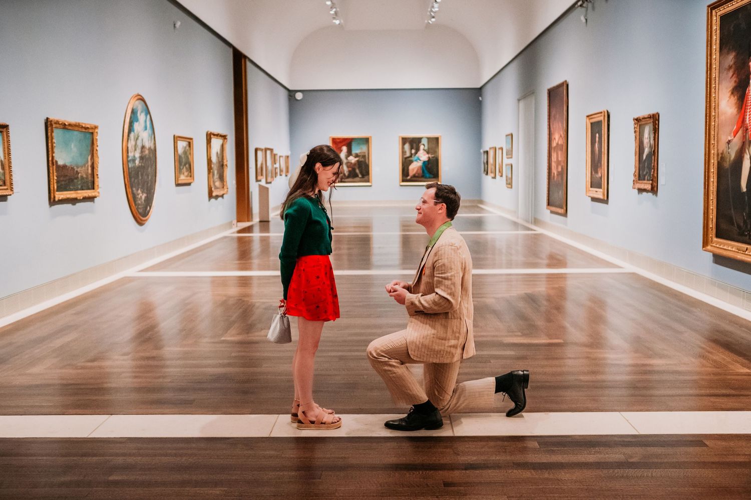 A romantic marriage proposal takes place in a spacious art gallery with wooden floors and paintings on white walls. MFAH