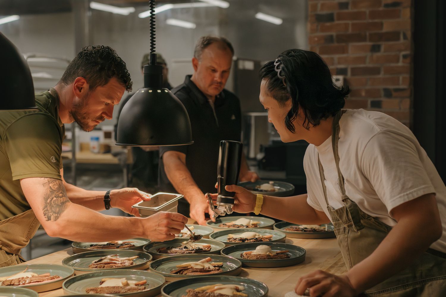 Chefs working together plating dishes in industrial restaurant kitchen with pendant lamps.