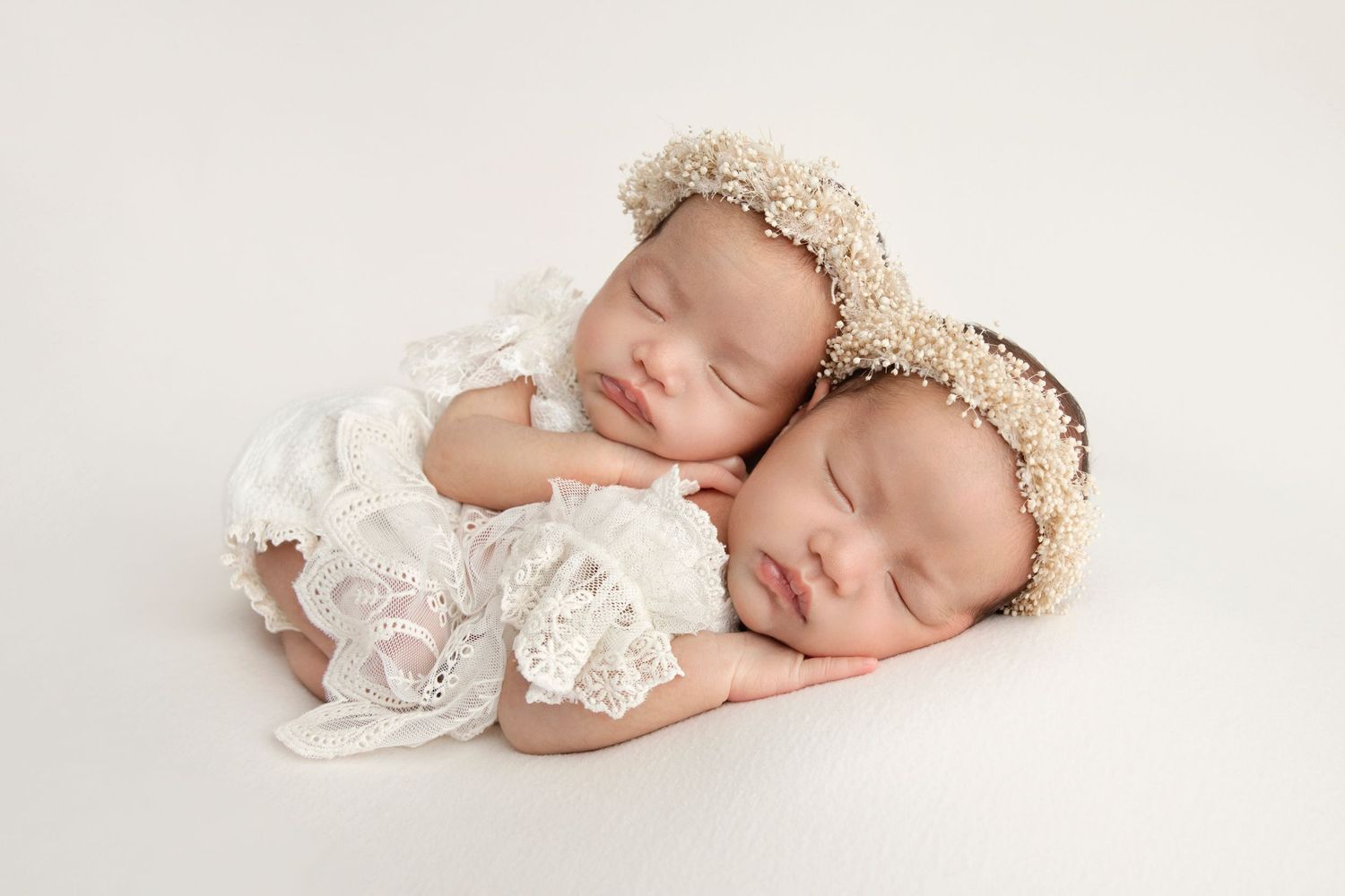 Two sleeping newborn babies wearing lace outfits and floral headbands laying together on a cream background.