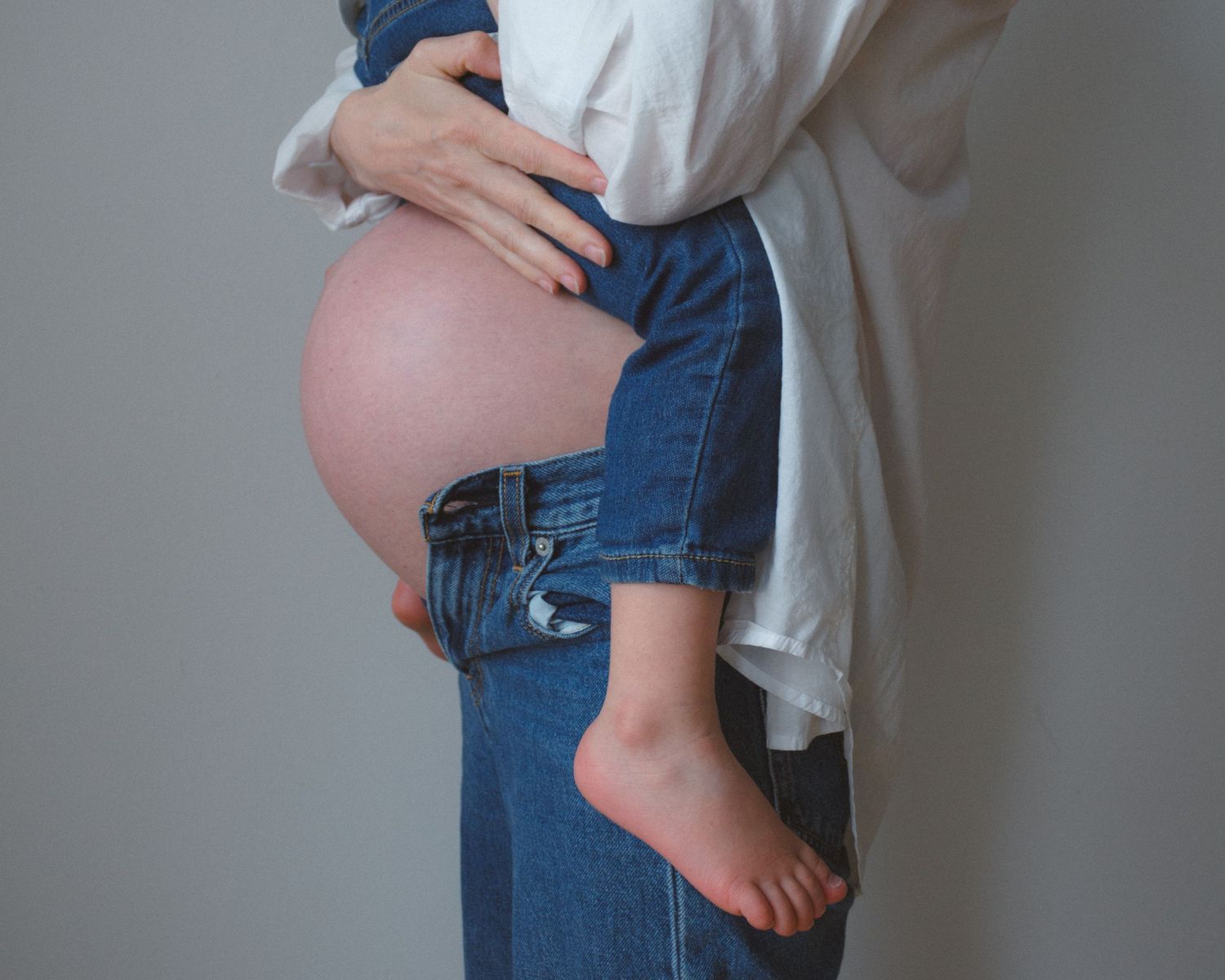Pregnant belly popping out of blue jeans while the mom is holding her toddler in a matching outfit on top of her belly for a cuddle.