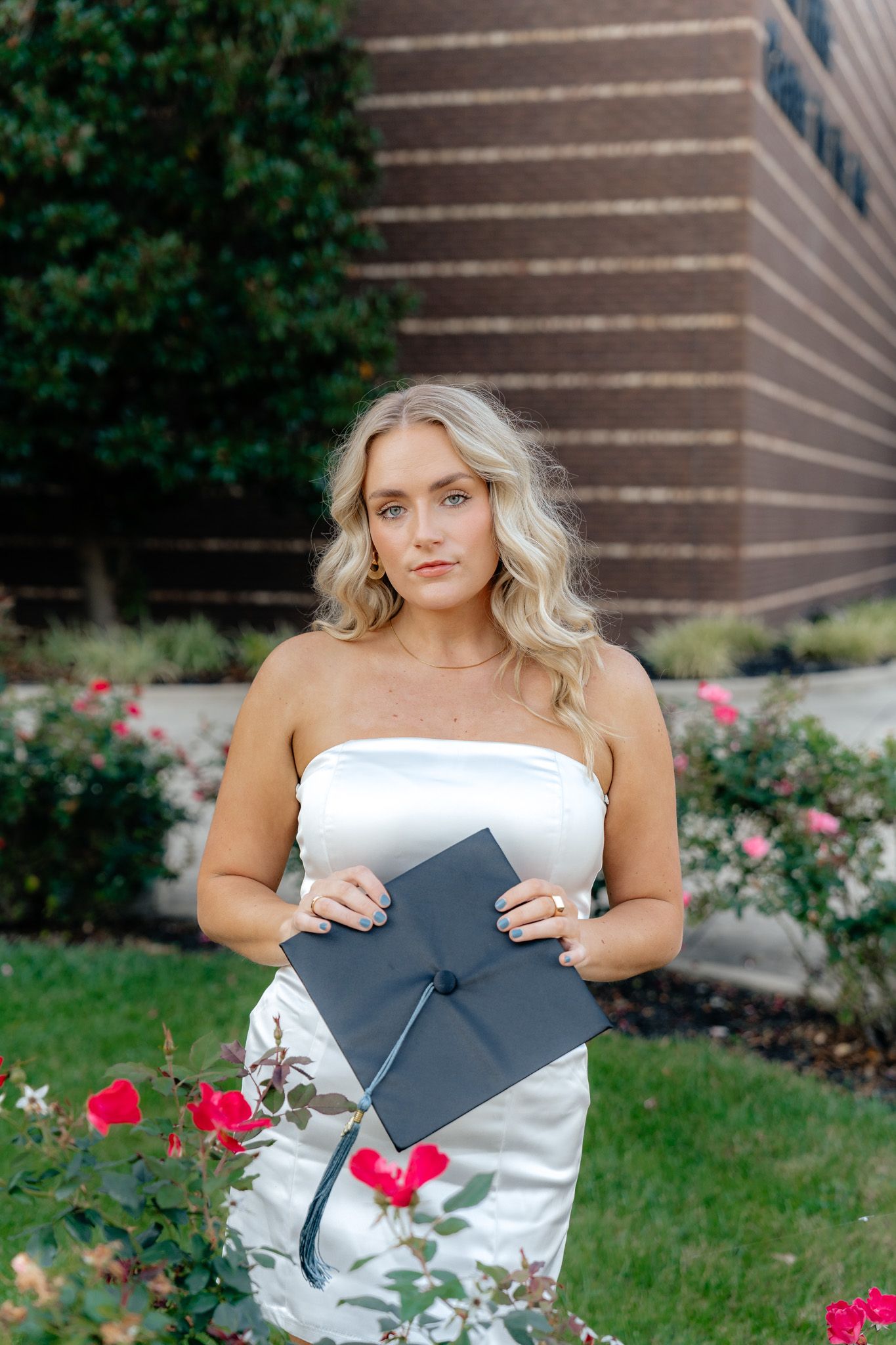 Graduate in white dress holding diploma and posing near blooming rose bushes.