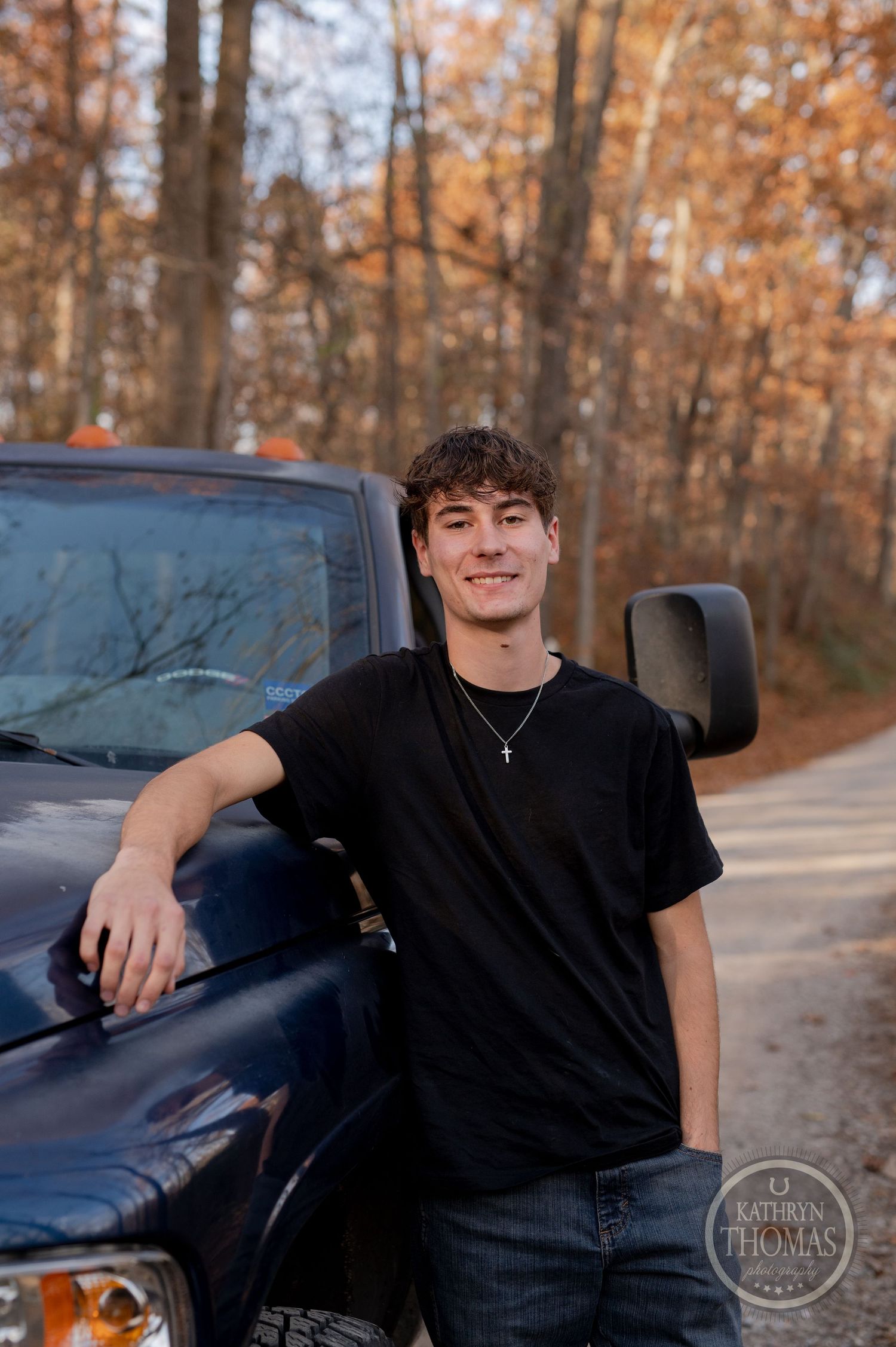 A casual portrait of someone leaning against a black Dodge Ram with autumn trees behind.