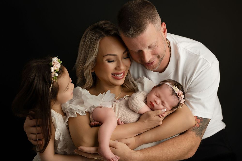 A sleeping newborn baby wearing a headband is held lovingly by parents in a professional studio portrait.