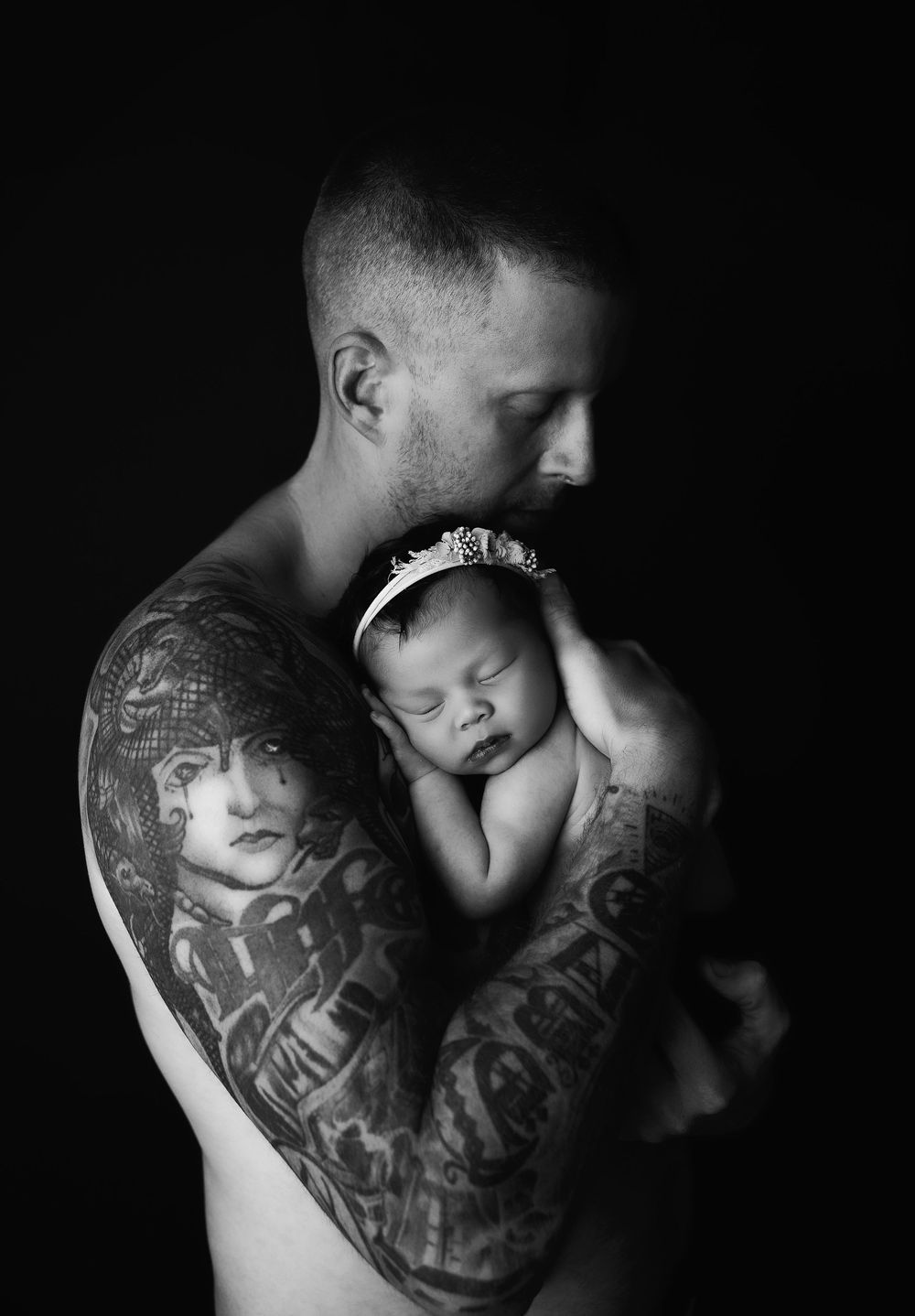A black and white portrait of a tattooed person holding a sleeping newborn baby against their bare chest.