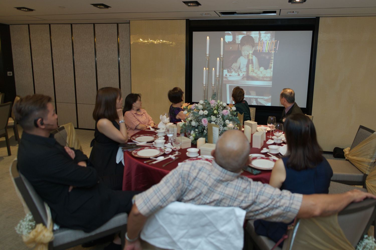 Wedding guests gather around a table watching a presentation on a projector screen in a banquet room.