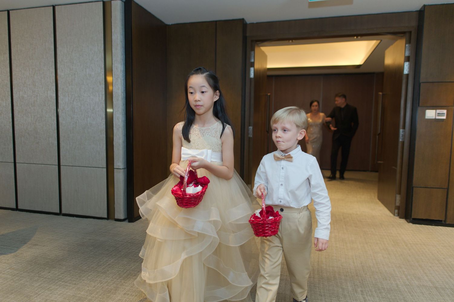 Young attendants in formal wear walking down hotel hallway carrying red bouquets.