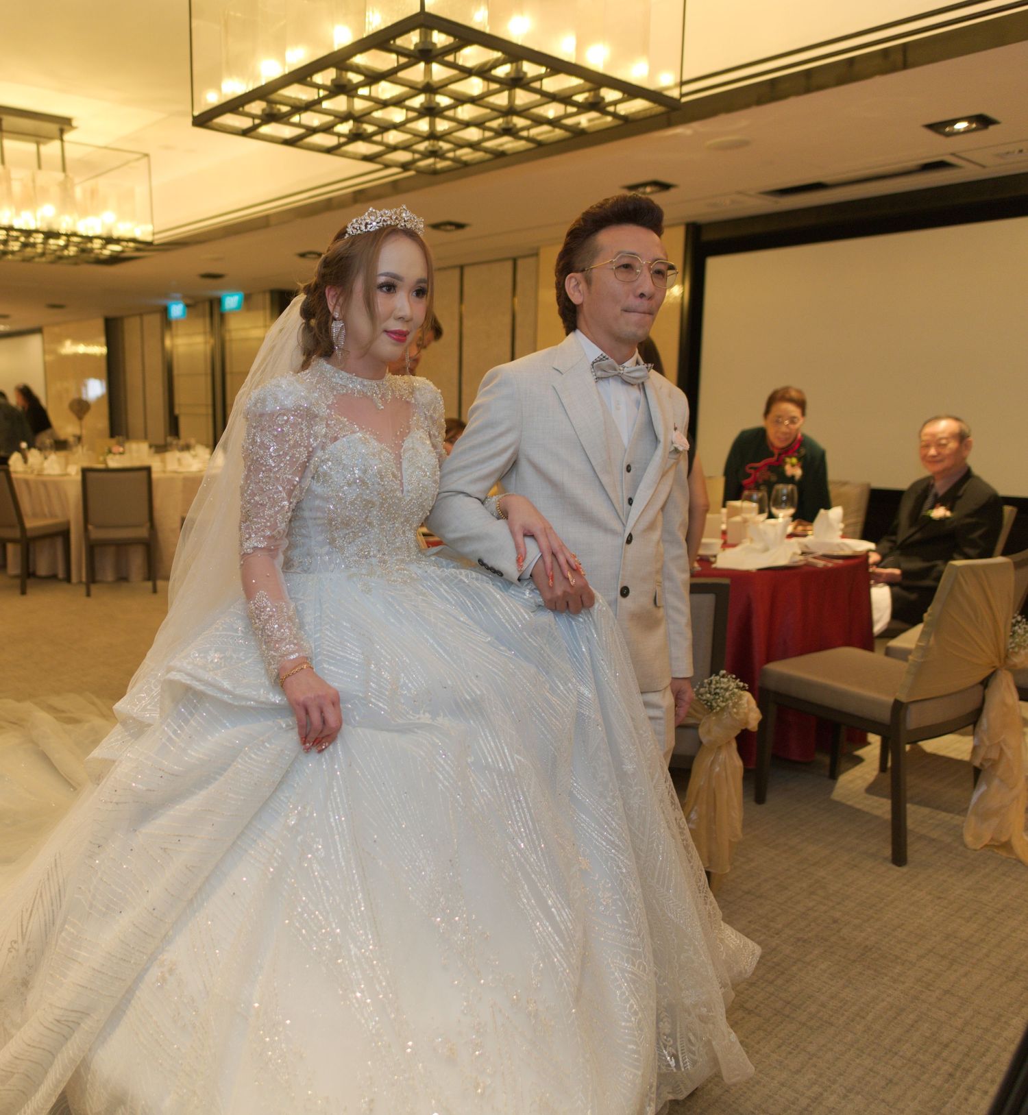 A couple in a grand banquet hall wearing elegant wedding attire, with a white lace gown and light-colored suit.