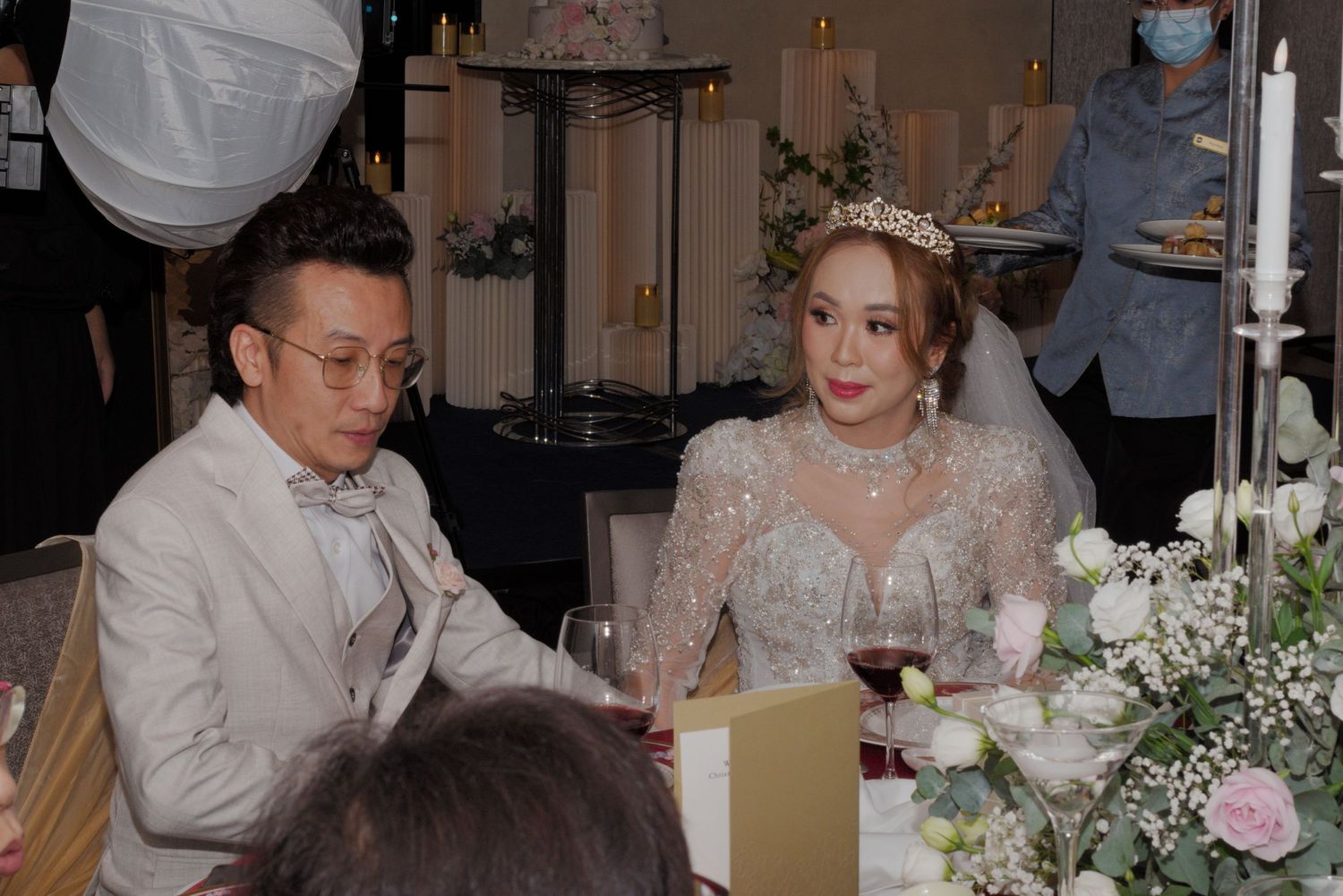 Couple sits at an elegantly decorated wedding reception table with white floral centerpieces and ambient lighting.