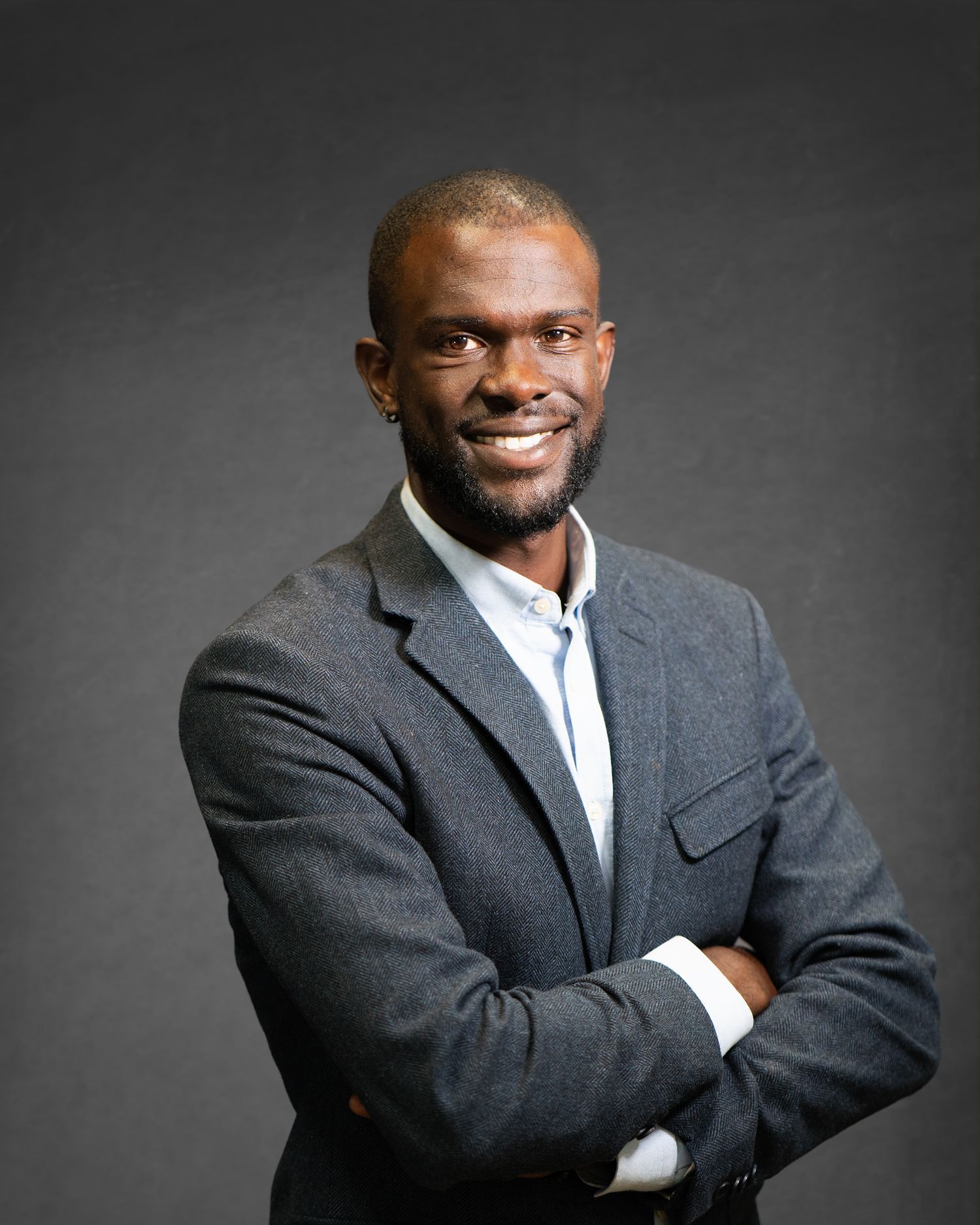 Professional headshot of someone in a grey suit jacket and white button-up shirt against a dark background.